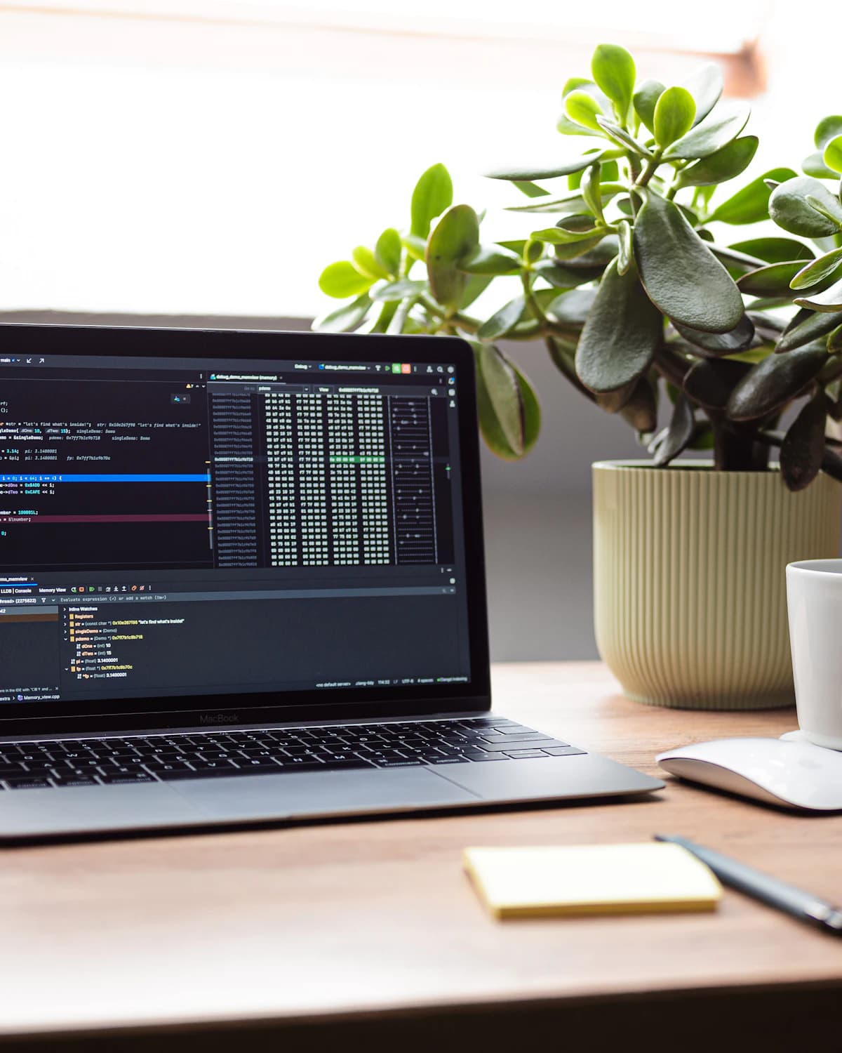 A laptop showing code on a wooden desk beside a plant and a coffee cup