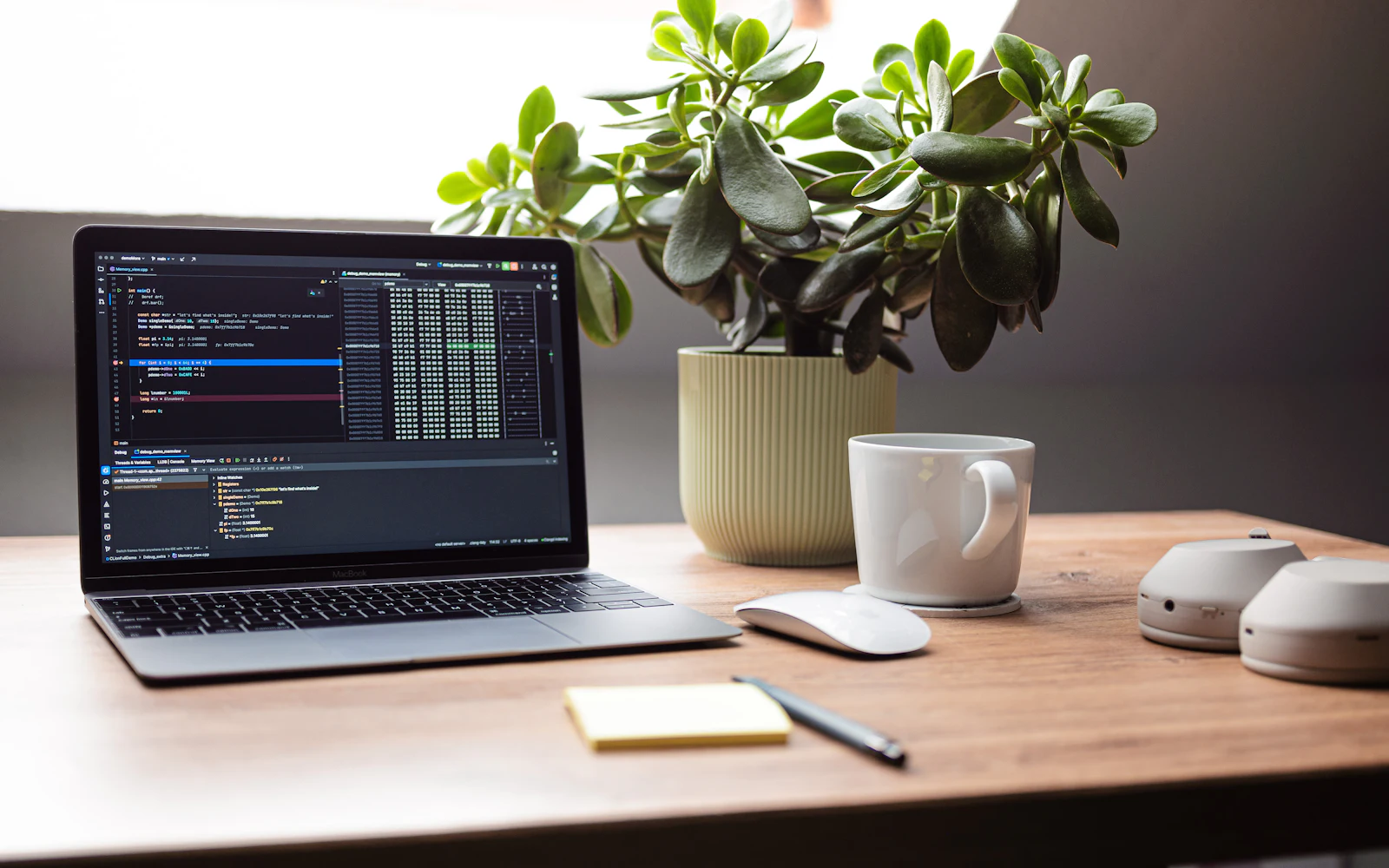 A laptop showing code on a wooden desk beside a plant and a coffee cup